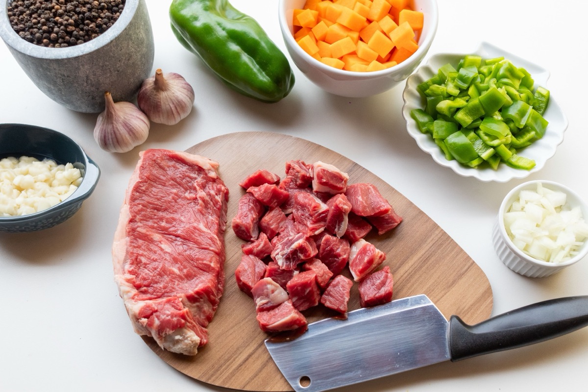 A beef steak and diced stewing beef on a wooden board, surrounded by prepared vegetables