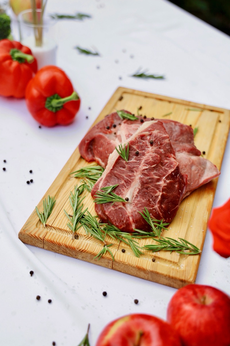 A marbled raw beef steak on a wooden board with rosemary and peppers
