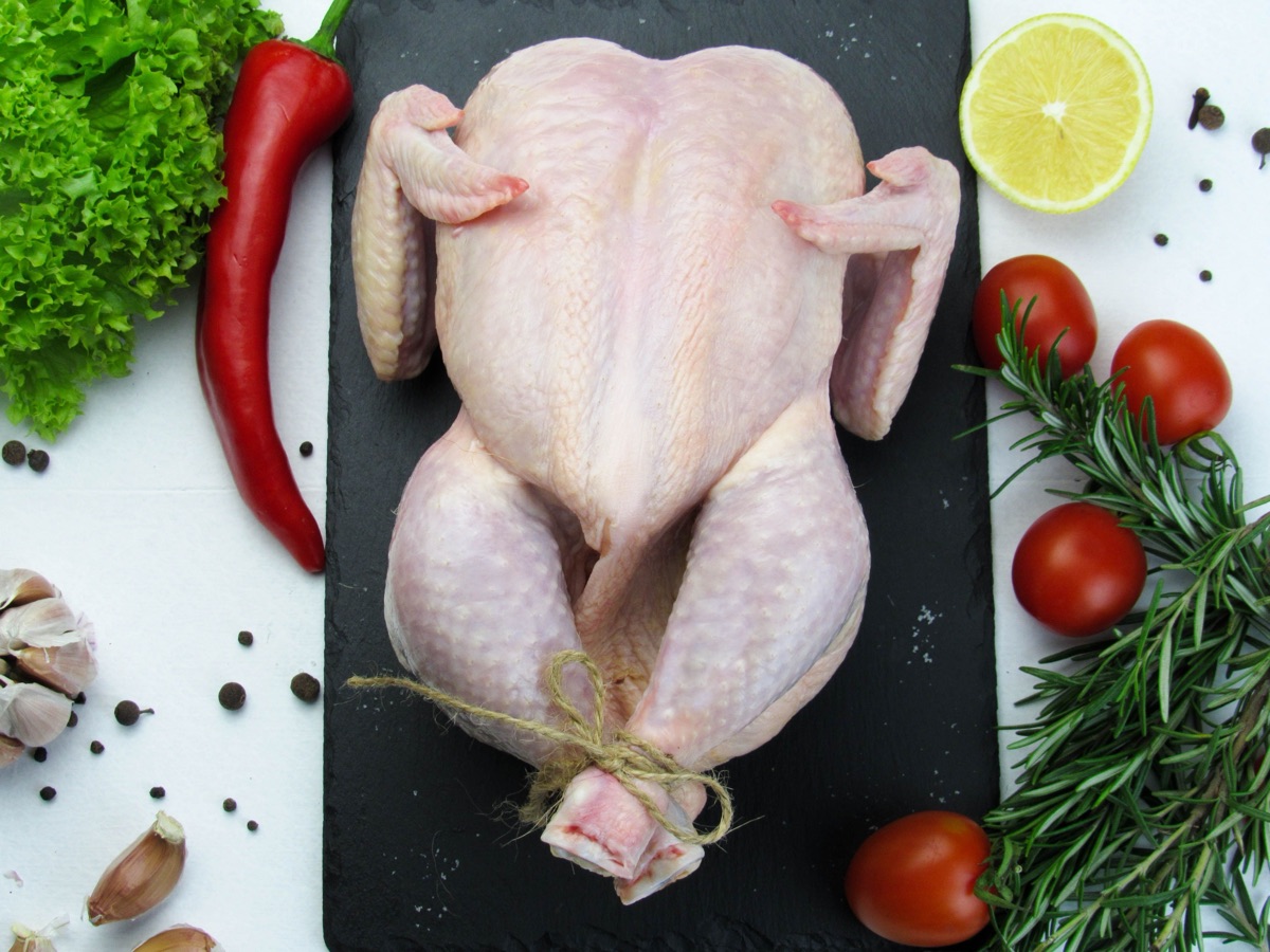 A whole raw chicken on a slate board surrounded by fresh vegetables and herbs